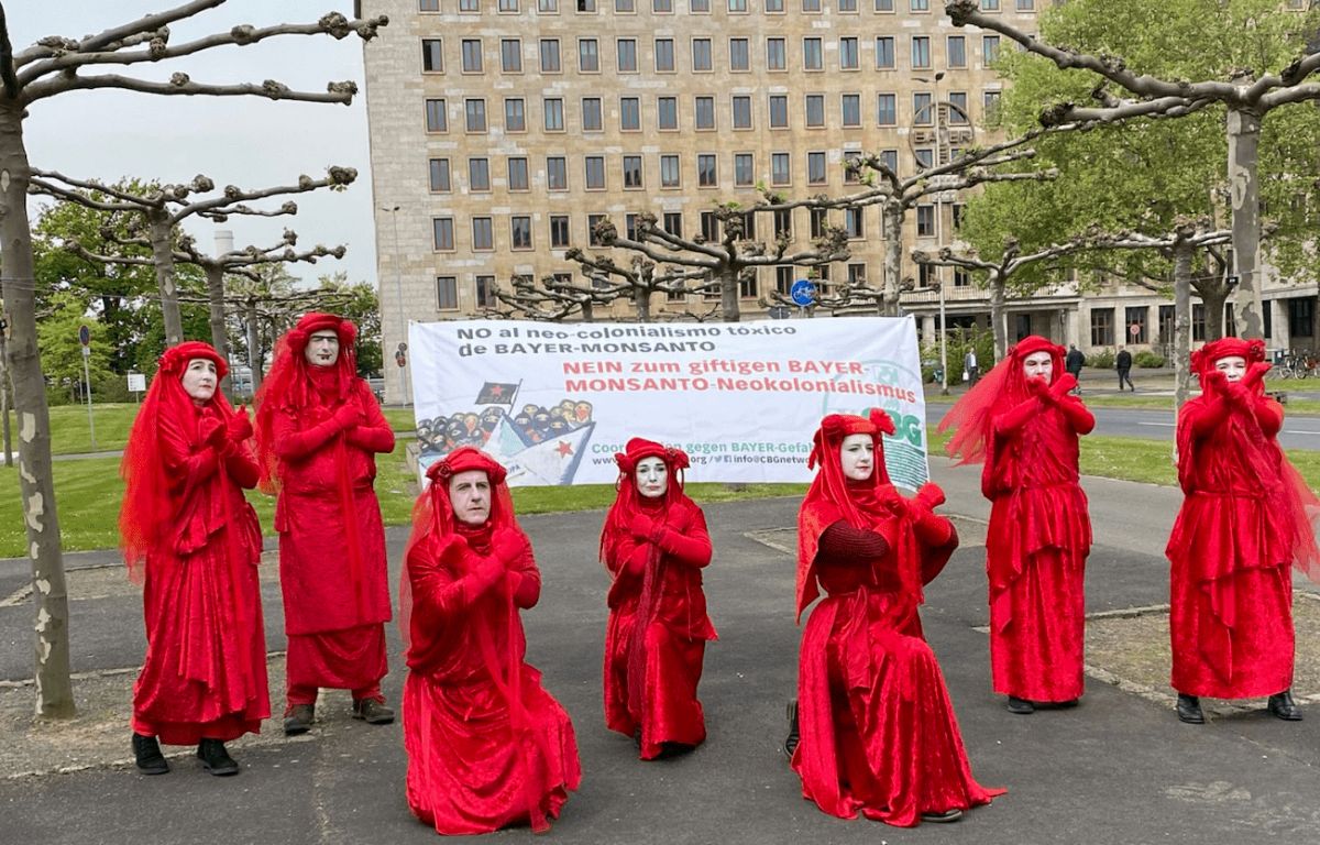 A group of people dressed in red costumes in front of a big banner near Bayer's offices protesting against Bayer.
