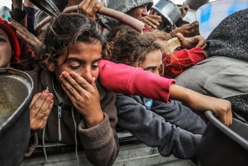 Children in in Al-Mawasi, southern Gaza, press forward in a crowded scene, some holding empty pots, their faces marked by distress and urgency.