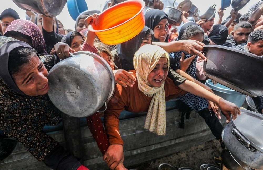 Children in in Al-Mawasi, southern Gaza, press forward in a crowded scene, some holding empty pots, their faces marked by distress and urgency.