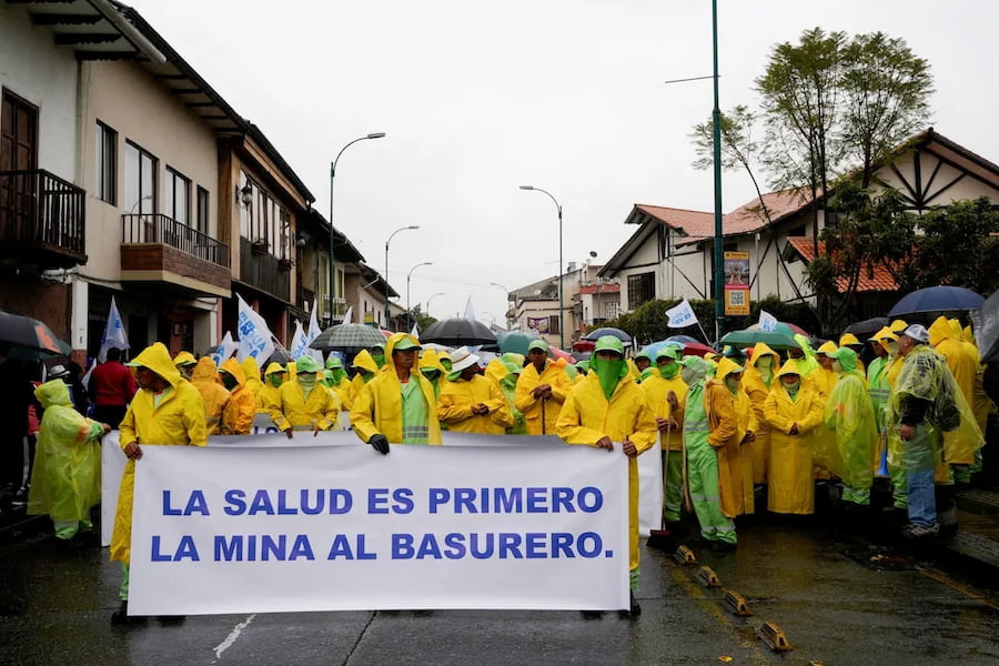 Protesters dressed in yellow coats stand on the street holding a banner that reads, " La Salud Es Primero, La Mina Al Basurero." They are protesting the Loma Larga gold mining project.