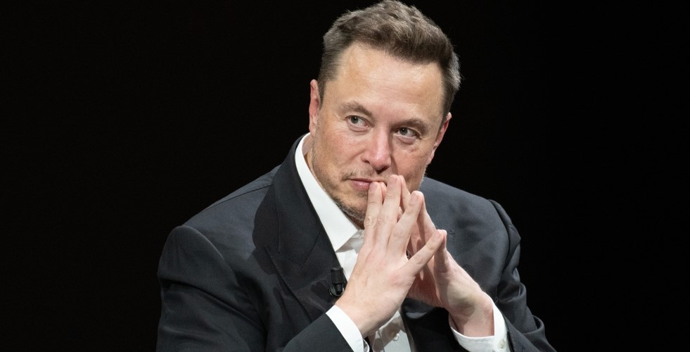 Elon Musk during an interview in 2023. He is wearing a white shirt and black suit and has his hands clasped together so that his fingertips are touching.