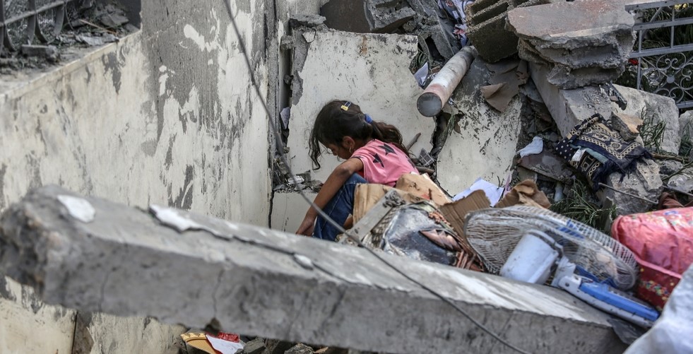 A young girl sits amid the shattered concrete and debris of a collapsed building, surrounded by fragments of everyday life.