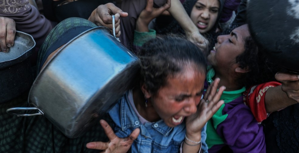 Women crowd together, trying to get meals from the Rafah charity kitchen (Tekka). The image captures pain and struggle as women and children push through with pots and pans in hand, faces tense with fear and exhaustion.