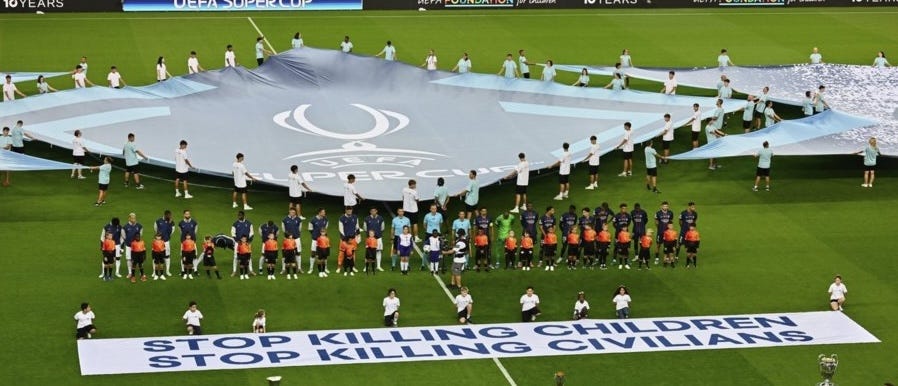 Football players at a UEFA-sponsored game line up with banners that say "Stop Killing Children" and "Stop Killing Civilians"